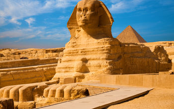 Full length body profile of Great Sphinx including head, feet with great pyramid of Menkaure in background on a clear, blue sky day in Giza, Egypt empty with no people. (photo via pius99 / iStock / Getty Images Plus)