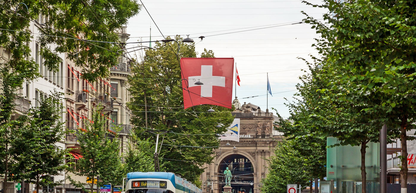 Image: PHOTO: Shopping promenade called Bahnhofstrasse in the city of Zurich. Tram / train with swiss flag in front. (photo via aldorado10 / iStock / Getty Images Plus)