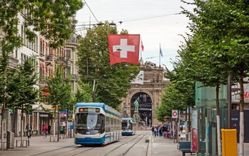 Zurich, Switzerland - June 10, 2017: Shopping promenade called Bahnhofstrasse, inner city of Zurich. Tram / train with swiss flag in front. (photo via aldorado10 / iStock / Getty Images Plus)