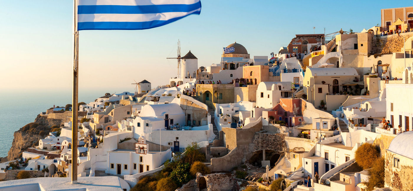 Image: The Greek flag flying at sunset in the town of Oia in Santorini, Greece. (photo via iStock/Getty Images Plus/ChrisHepburn)