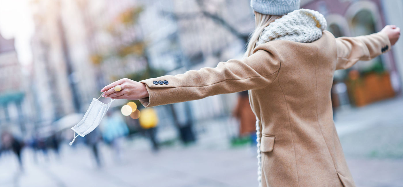 Image: Woman taking off face mask in the city. (photo via macniak / iStock / Getty Images Plus)