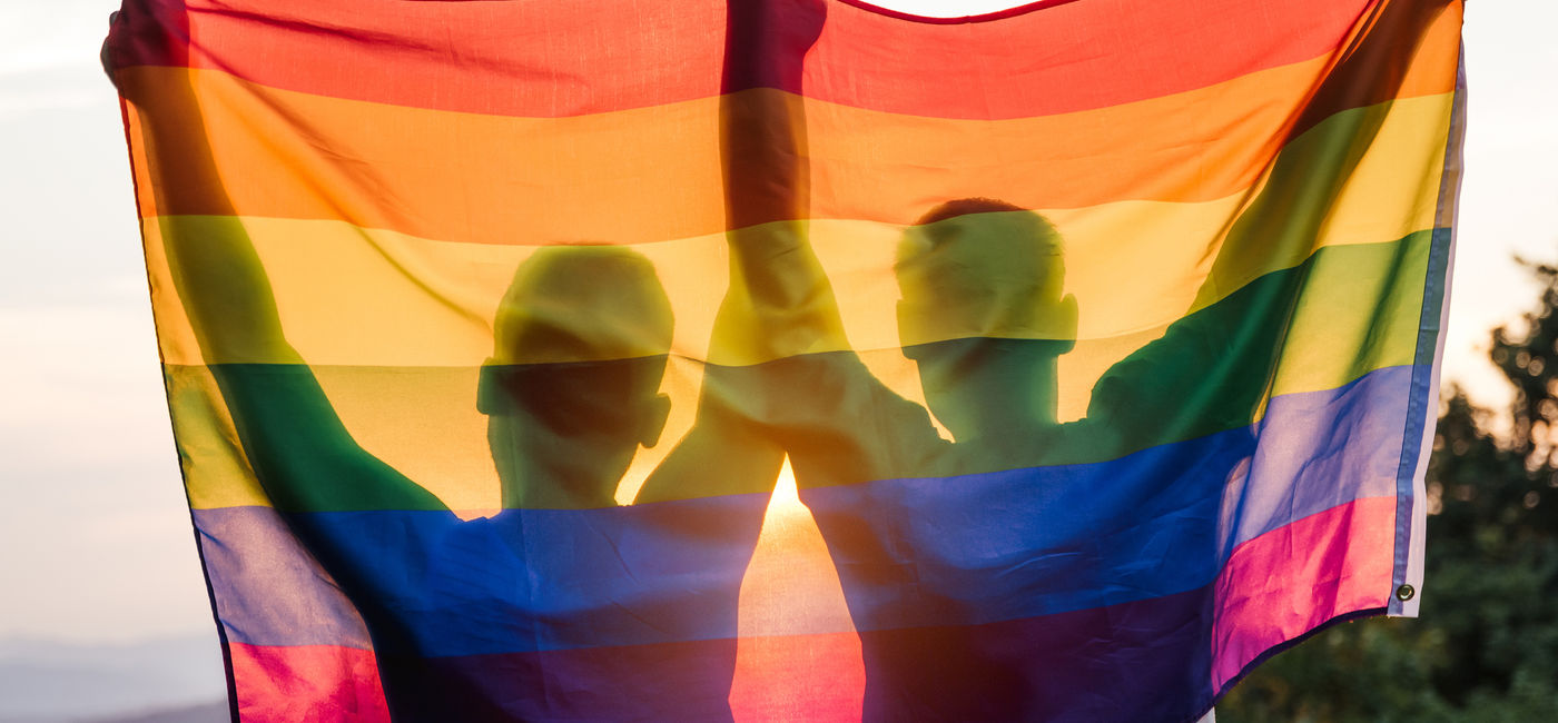 Image: A gay couple holding up a rainbow flag at sunset. (Photo via iStock/Getty Images E+/D-Keine)