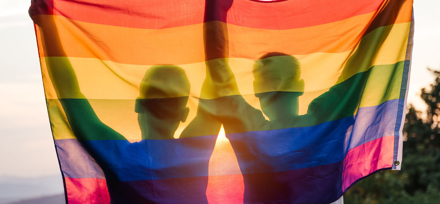 Photo: A gay couple holding up a rainbow flag at sunset. (Photo via iStock/Getty Images E+/D-Keine)