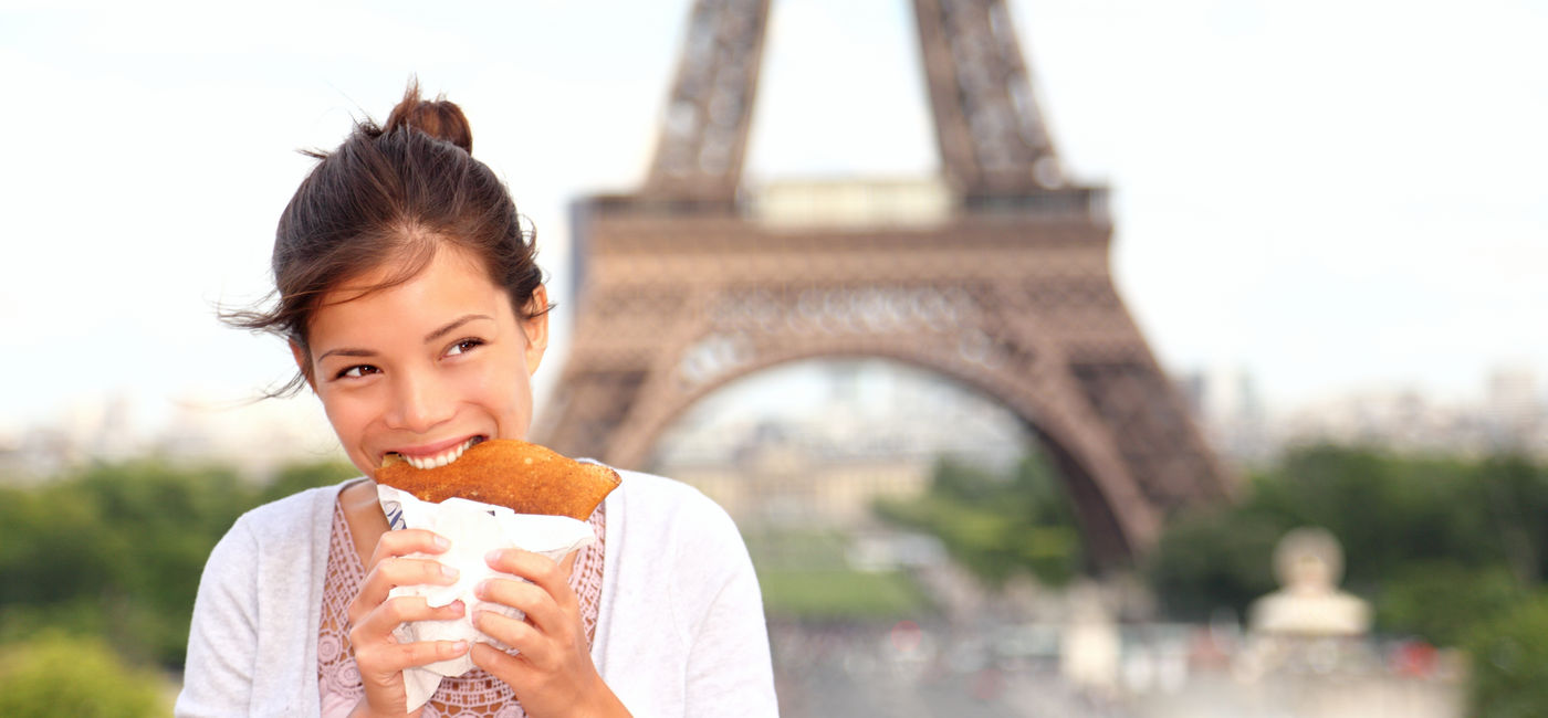 Photo: Woman enjoying street food in front of the Eiffel Tower in Paris, France. (photo via ariwasabi/iStock/Getty Images Plus)