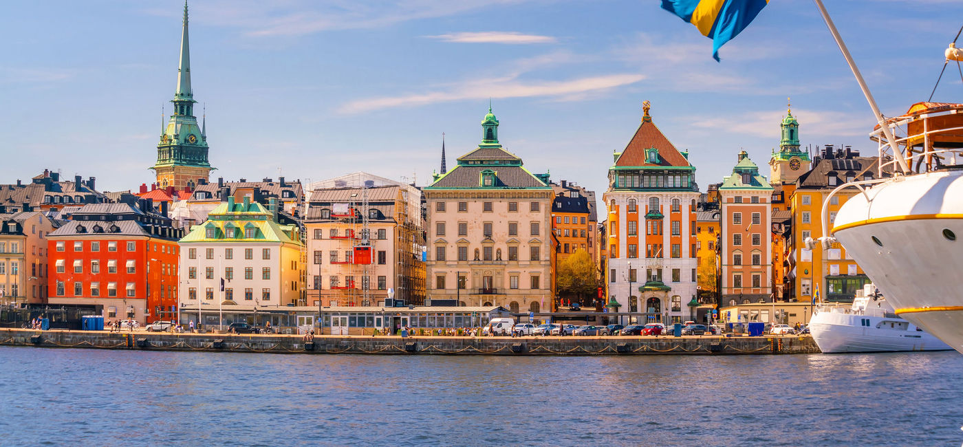 Image: Waterfront in Old Town Stockholm with Sweden's national flag in the foreground. (photo via iStock/Getty Images Plus/f11photo)