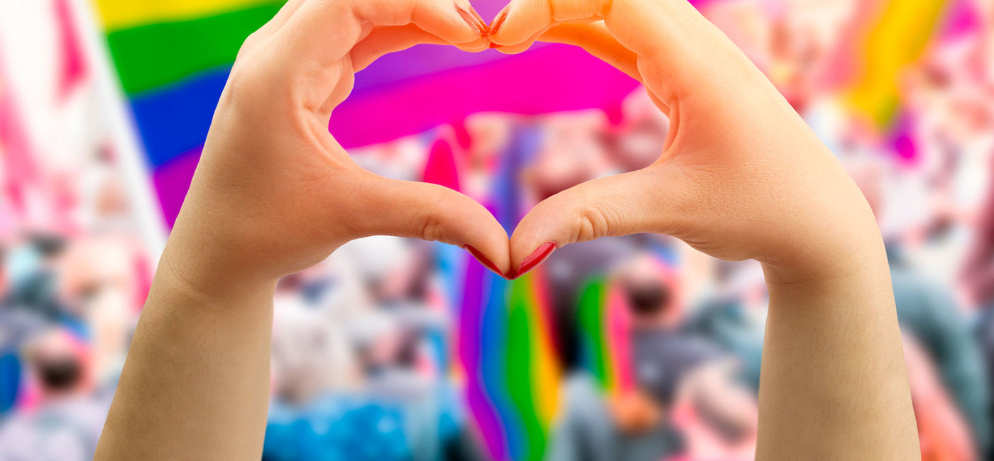 Image: Hands forming the shape of a heart in front of a rainbow flag flying at an LGBTQ+ Pride parade. (photo via iStock/Getty Images Plus/Cunaplus_M.Faba)