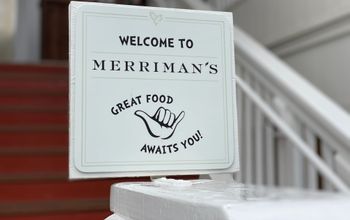Sign reading &#34;Welcome to Merriman&#39;s - Good Food Awaits You&#34; with a shaka hand gesture