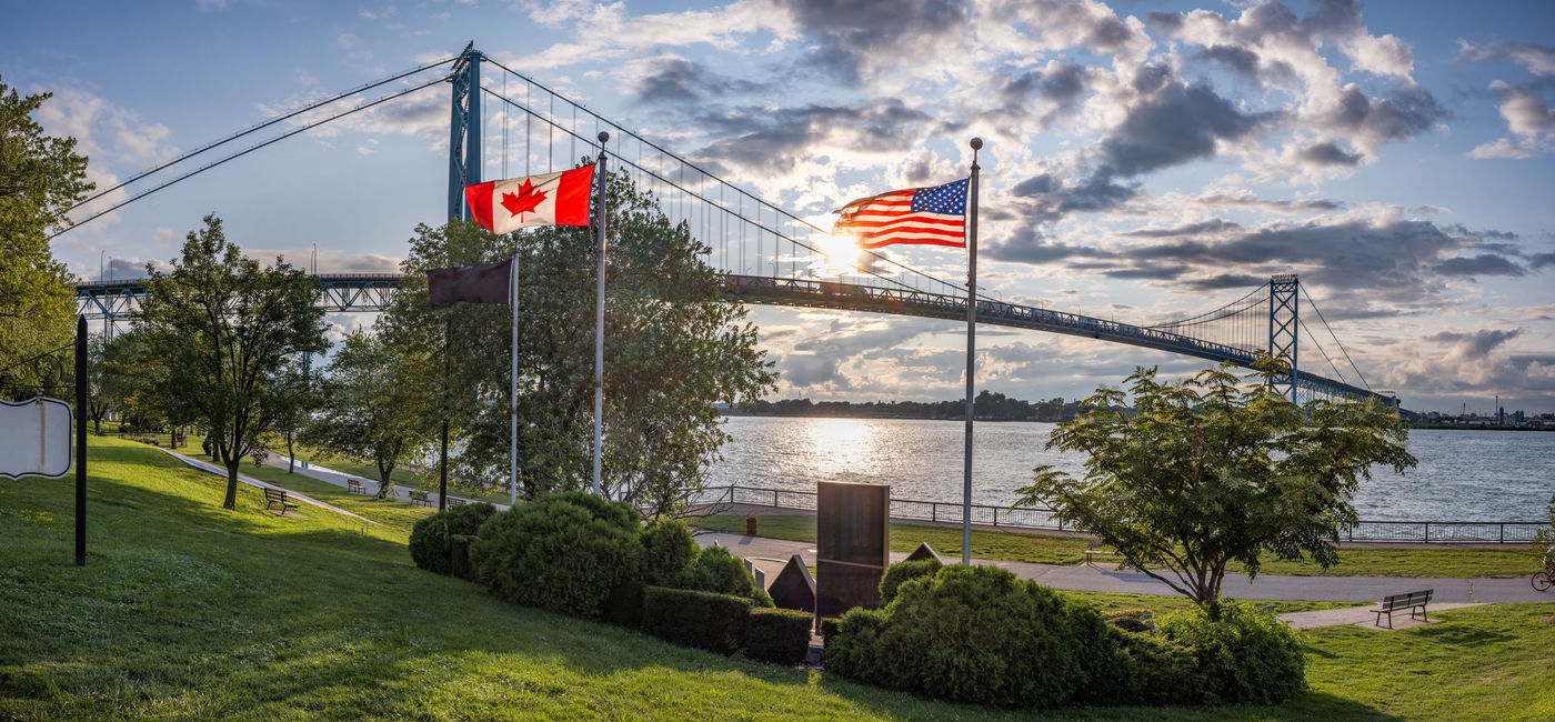 Image: The U.S. and Canadian flags flying in front of the Ambassador Bridge, which links Detroit, Michigan with Windsor, Ontario. (photo via iStock/Getty Images Plus/Steven_Kriemadis)