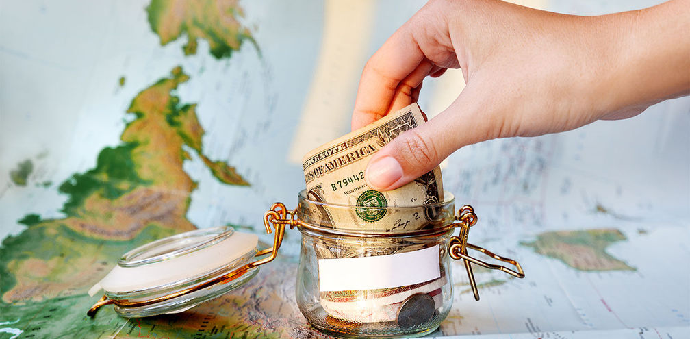 Collecting money for travel. Glass tin as moneybox with cash (Photo via Aksenovko / iStock / Getty Images Plus)