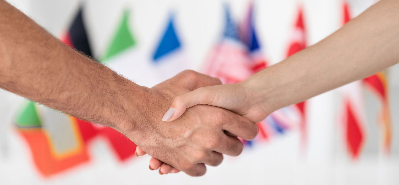 Image: People shaking hands with various national flags in the background. (Photo Credit: iStock/Getty Images Plus/Prostock-Studio)