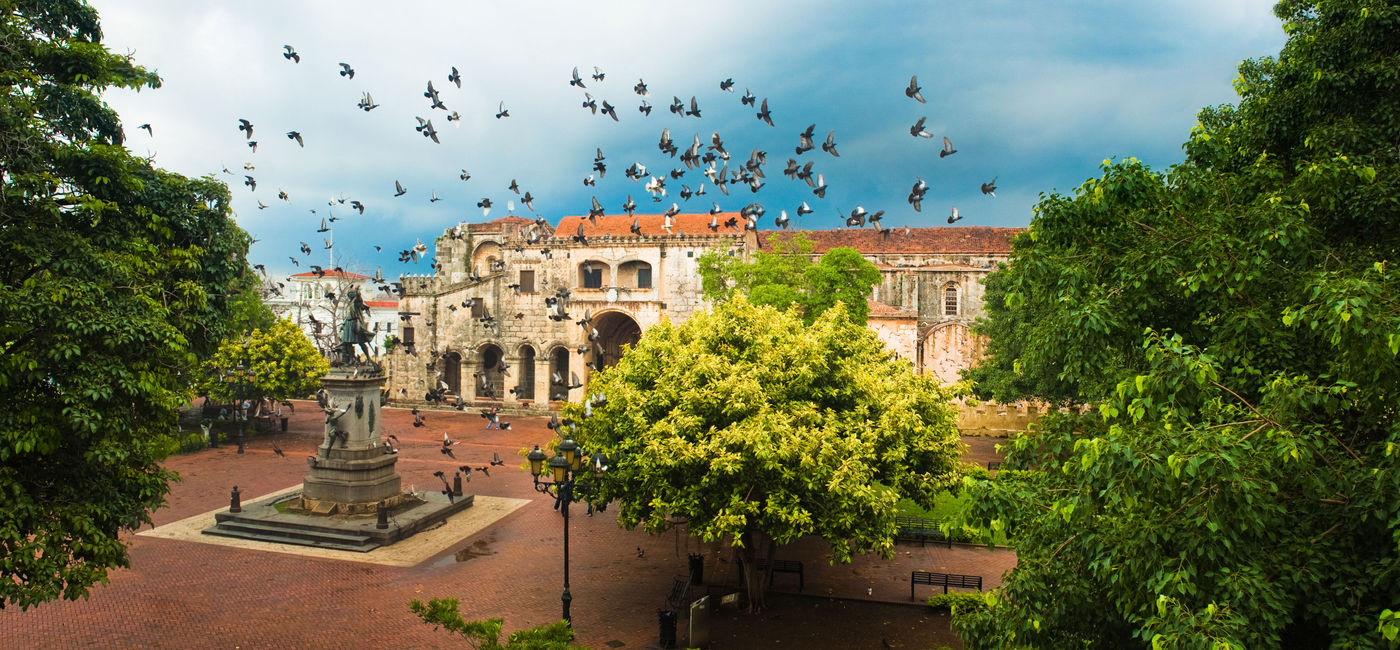 Image: Doves flying over the main square with Columbus statue, Santo Domingo, Dominican Republic. (photo via 3dan3 / iStock / Getty Images Plus)