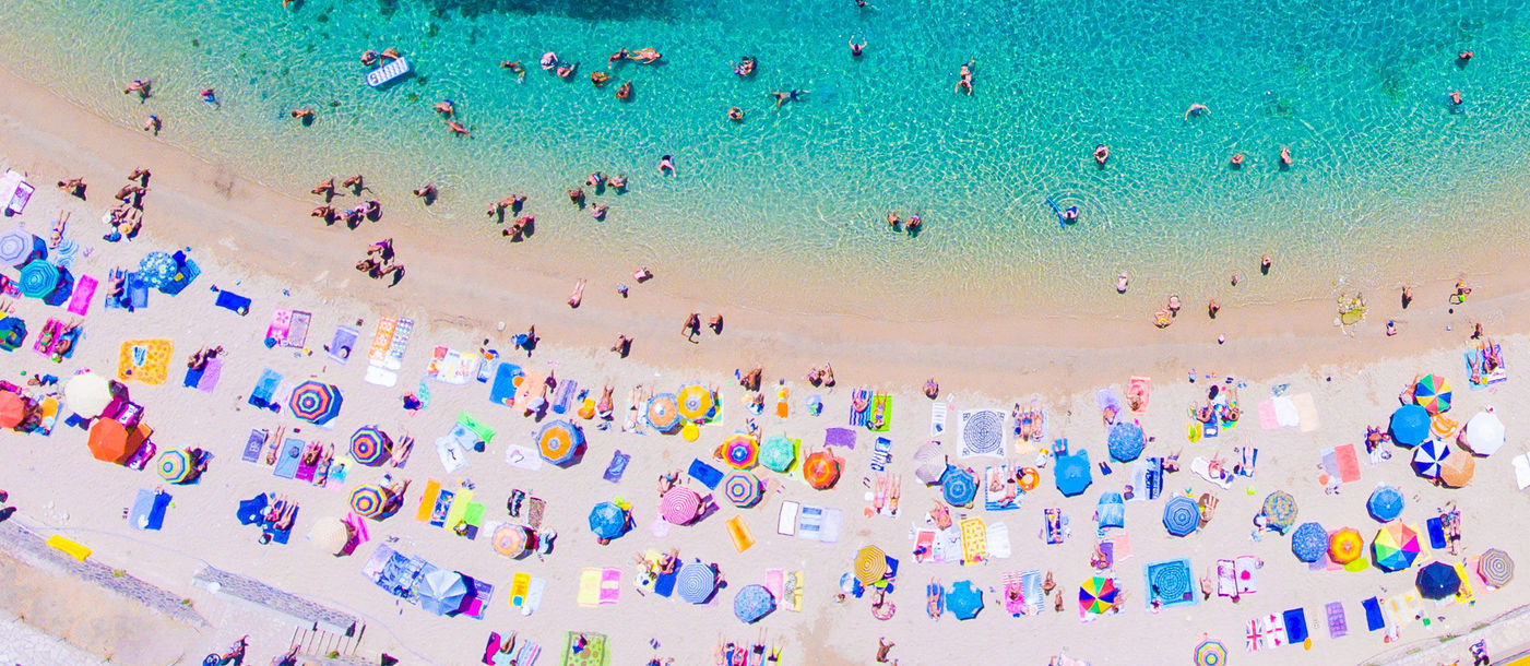 People bathing in the sun at the beach, aerial view with clear blue water and sandy beach. Corfu island Kerkyra, Greece (photo via CalinStan/iStock/Getty Images Plus)