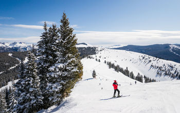 Coniferous forest covered by snow with skiing slopes in the foreground and Rocky Mountains in the background. This photograph was taken in Vail, Colorado. (photo via miralex / iStock / Getty Images Plus)