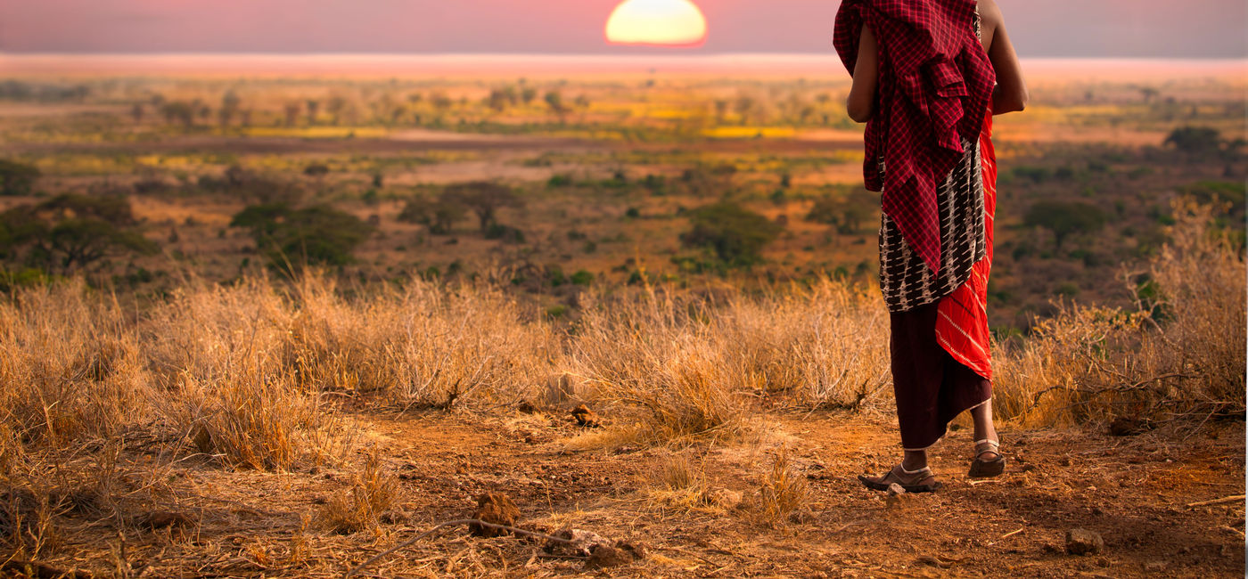 Image: Masai man, wearing traditional blankets, overlooks Serengeti in Tanzania as the colorful sunset fills the sky. Wild grass in the forground. (photo via jocrebbin/iStock/Getty Inages Plus)