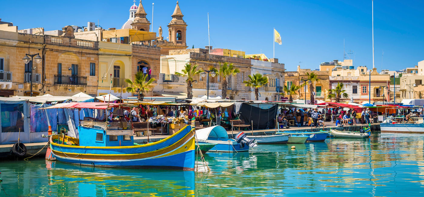 Image: Marsaxlokk market in Malta with traditional Luzzu fishing boats on a beautiful summer day. (photo via ZoltanGabor / iStock / Getty Images Plus)