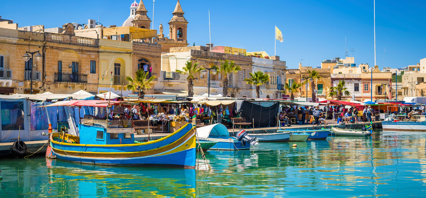 Image: Marsaxlokk market in Malta with traditional Luzzu fishing boats on a beautiful summer day. (photo via ZoltanGabor / iStock / Getty Images Plus)