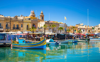 Malta - Marsaxlokk market with traditional Luzzu fishing boats on a beautiful summer day withblue sky and green sea (photo via ZoltanGabor / iStock / Getty Images Plus)
