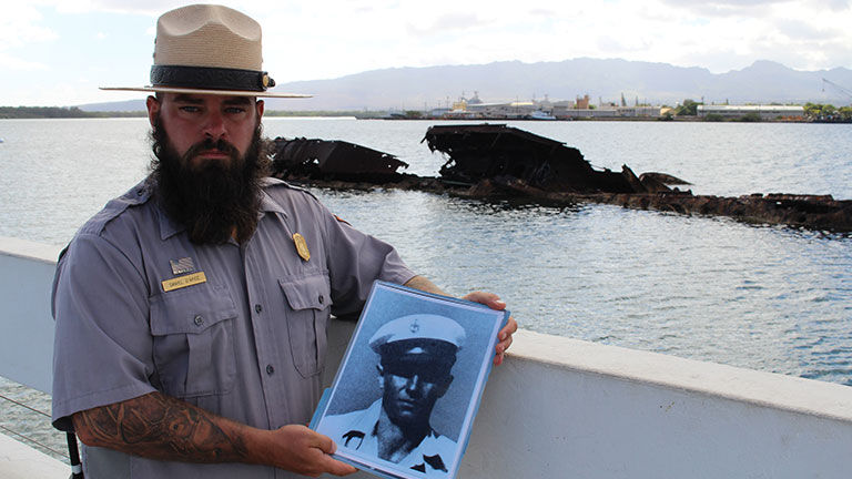 National Park Service Ranger Daniel D’Apice shows a photo of a USS Utah crew member at the USS Utah Memorial, with the wreckage of the partially submerged vessel in the background.