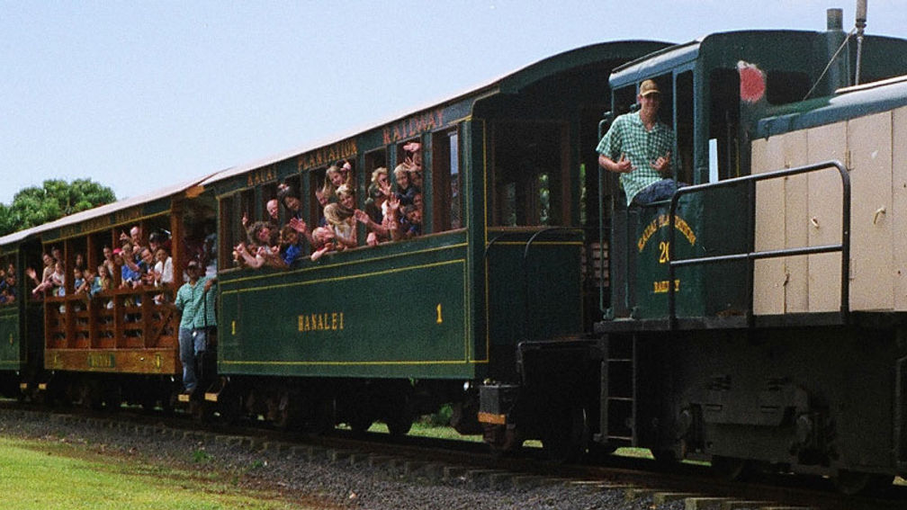 A vintage train on the Kauai Plantation Railway chugs through a field of tropical trees on Kauai. Passengers enjoy the scenic ride, with lush green mountains potentially visible in the distance. [Courtesy Kauai Plantation Railway]
