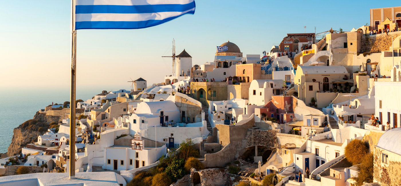 Image: The Greek flag flying at sunset in the town of Oia in Santorini, Greece. (photo via iStock/Getty Images Plus/ChrisHepburn)