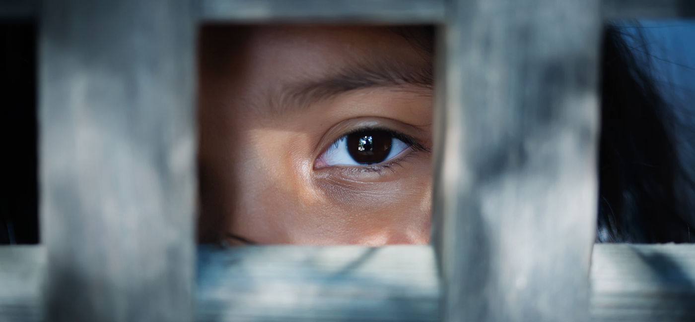 Image: A child staring out from behind wooden slats. (photo via iStock/Getty Images Plus/mmg1design)