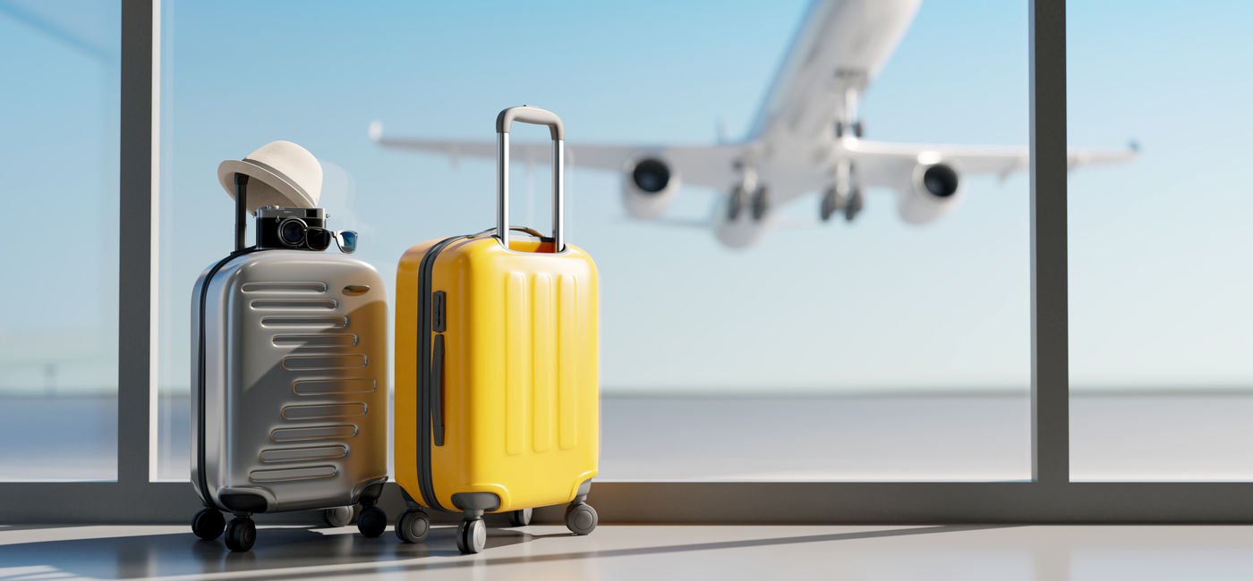 Image: Luggage sitting at the airport with a plane taking off in the background. (Photo Credit: Adobe Stock/aanbetta)