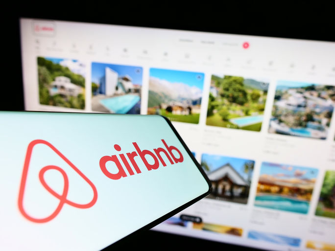 Airbnb logo displayed on a smartphone in front of a listings webpage.