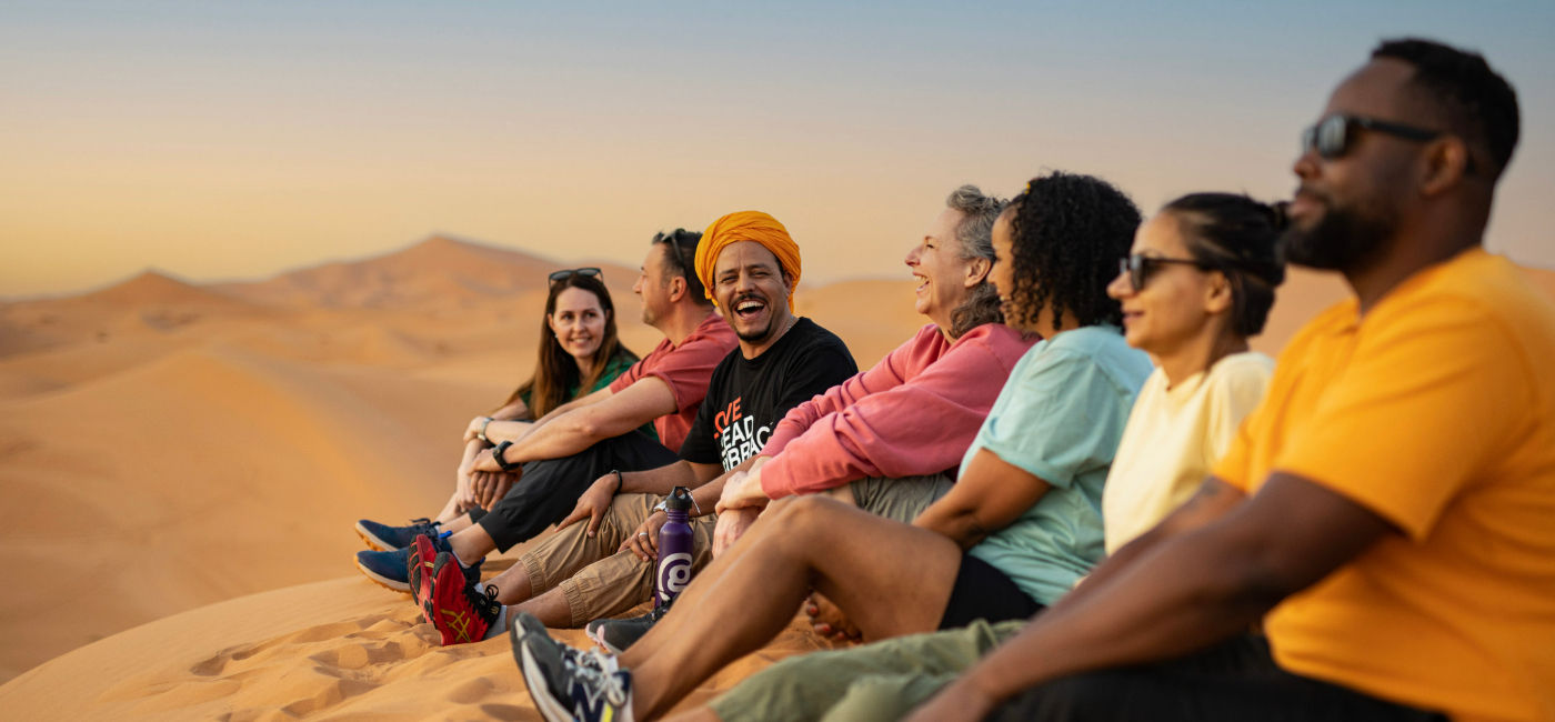 Image: A G Adventures group CEO and travellers sit in the sand dunes in Morocco. (Photo Credit: G Adventures)