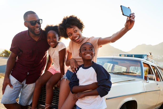 Family posing for selfie next to car packed for road trip.