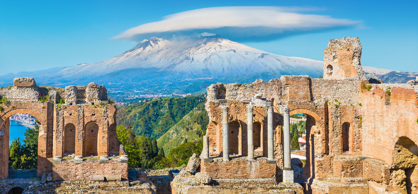 Image: Mount Etna towers above ancient theatre ruins in Taormina, Sicily. (Photo Credit: IgorZh / Adobe Stock)