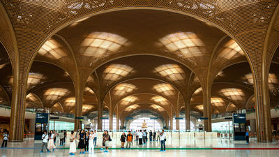 The arrival hall features a soaring tree-canopy-inspired ceiling, warm neutral tones, indoor greenery, and a grid-shell roof that filters natural light throughout the space.