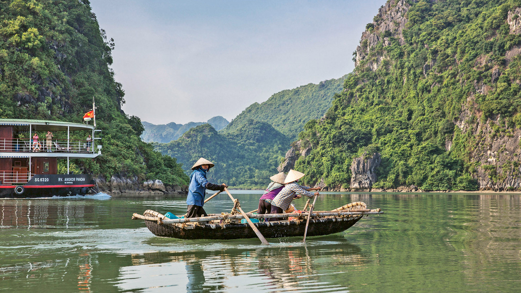 The Angkor Pandaw cruises past a rustic boat and impressive karst formations in Ha Long Bay, Vietnam.