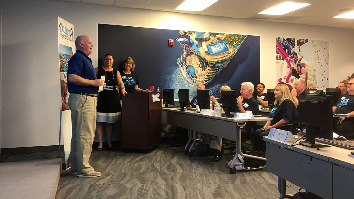 A new CruiseOne/Cruises Inc/DreamVacations franchisee is recognized for training achievement in a room decorated with murals of Royal Caribbean International ships.