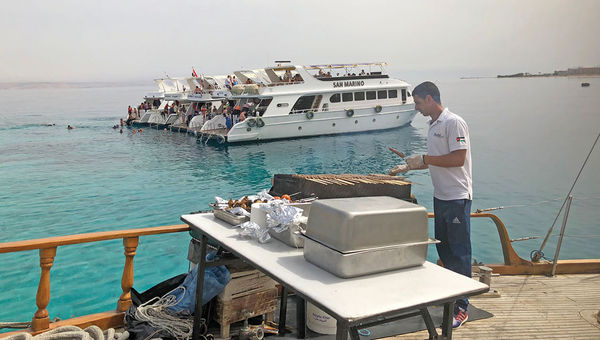 A deckhand prepares a barbecue for passengers on an excursion boat in the Gulf of Aqaba. In the background, dive boats are rafted up over a snorkeling site.