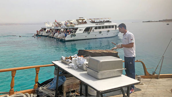 A deckhand prepares a barbecue for passengers on an excursion boat in the Gulf of Aqaba. In the background, dive boats are rafted up over a snorkeling site. A deckhand prepares a barbecue for passengers on an excursion boat in the Gulf of Aqaba. In the background, dive boats are rafted up over a snorkeling site.