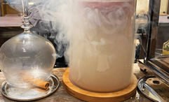 A burning cinnamon stick sits below a glass that will hold the Figsation drink while another drink called the Benevolent Dictator is smoked beside it.