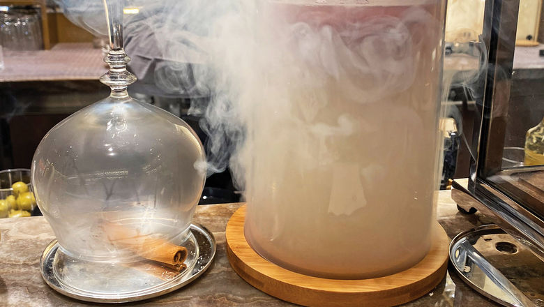 A burning cinnamon stick sits below a glass that will hold the Figsation drink while another drink called the Benevolent Dictator is smoked beside it.