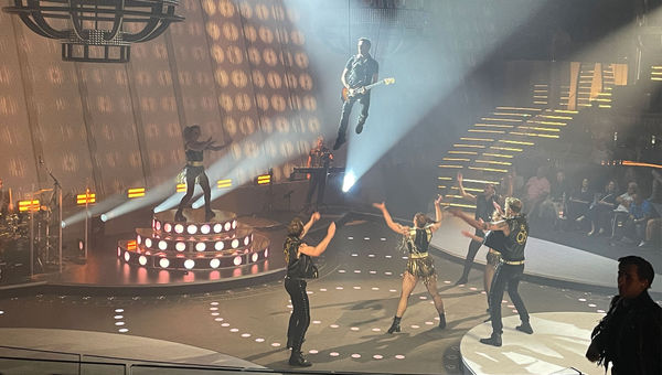 Dançarinos e cantores da "Queen Symphonic" se apresentam em um amplo palco e ao longo das escadas do Panorama Lounge, enquanto o guitarrista alça voo.