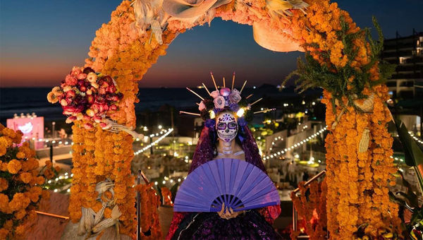 A woman in an elegant dress and skull face paint, called a Catrina, stands under an archway made of Mexican marigolds called cempasuchils. A woman in an elegant dress and skull face paint, called a Catrina, stands under an archway made of Mexican marigolds called cempasuchils.