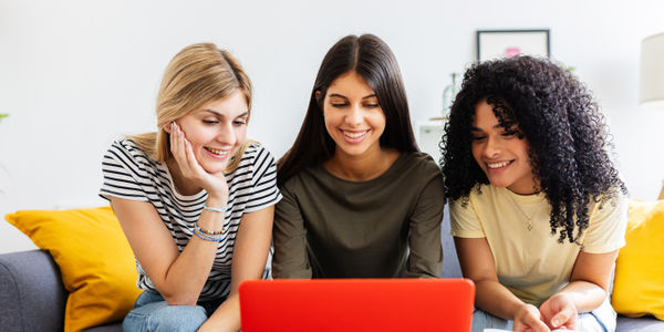 Group of young women planning travel together using laptop at home. Friends searching destinations and organizing trip details while enjoying time together indoors. Travel and lifestyle concept.