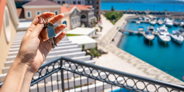 Hand holding a house key with a blurred seaside villa and marina in the background. Concept of buying coastal real estate, property investment and owning a vacation home