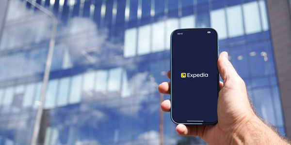 Expedia mobile travel booking application displayed on smartphone held in male hand near modern office building