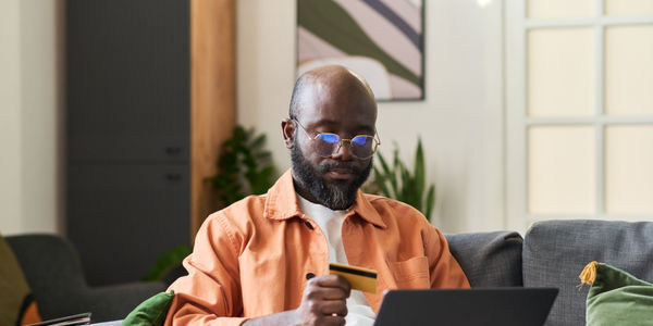 Medium shot of Black middle aged man holding credit card and using laptop while booking trip from home, planning travel purchase. Useful for tourism marketing, online payment