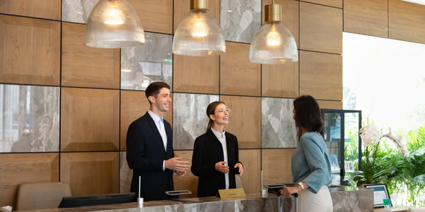 Female guest standing at hotel reception desk with suitcase during check-in, talking with receptionists, confirming reservation details. Hospitality, customer services concept for travel and tourism