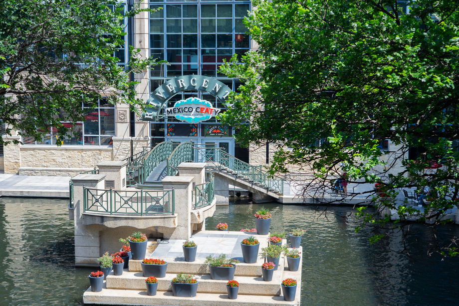 Floating stone platform with many potted flowers sits on a canal by a building and bridge.