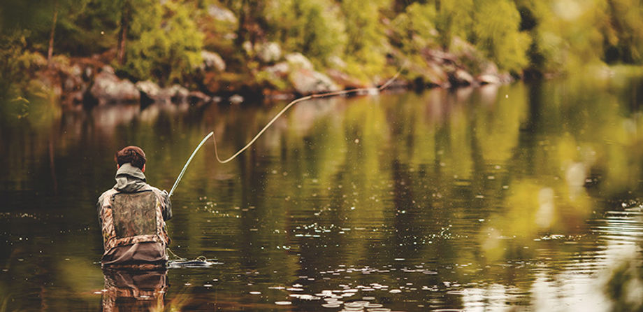A fisherman wearing camouflage stands waist-deep in a calm river and casts a line.