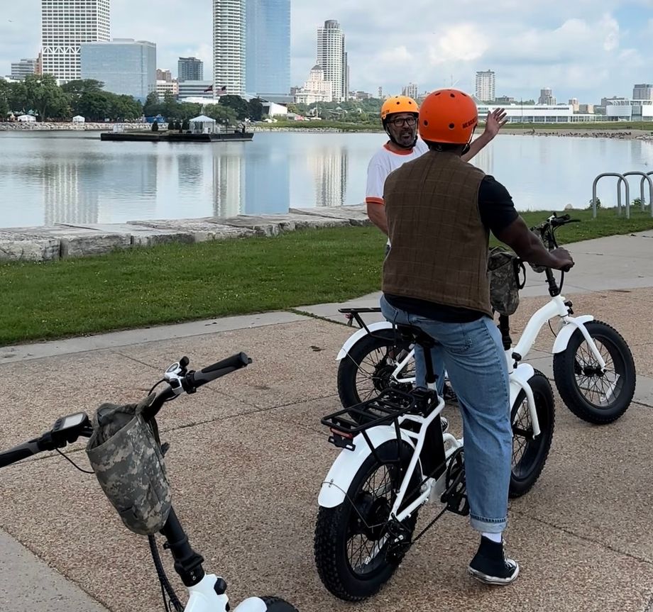 Two people wearing helmets chat beside white electric bikes by a riverfront park.