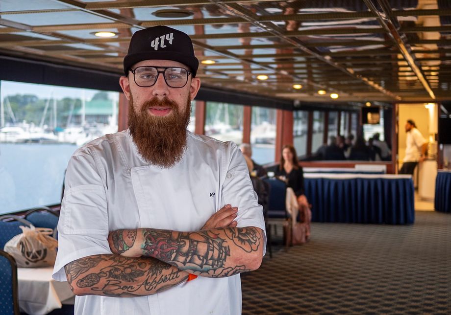 A bearded chef with glasses stands confidently on a boat deck.