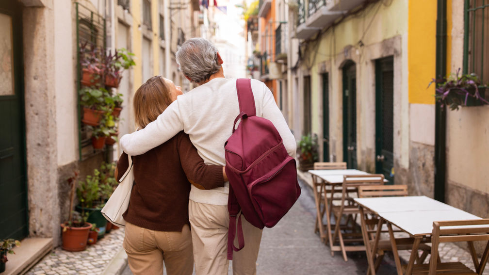 Vacation Tour Offer. Back View Of Senior Couple Hugging And Carrying Backpack Walking On Lisbon Street, Enjoying Sightseeing Outdoors. Travel And Tourism In Europe Concept