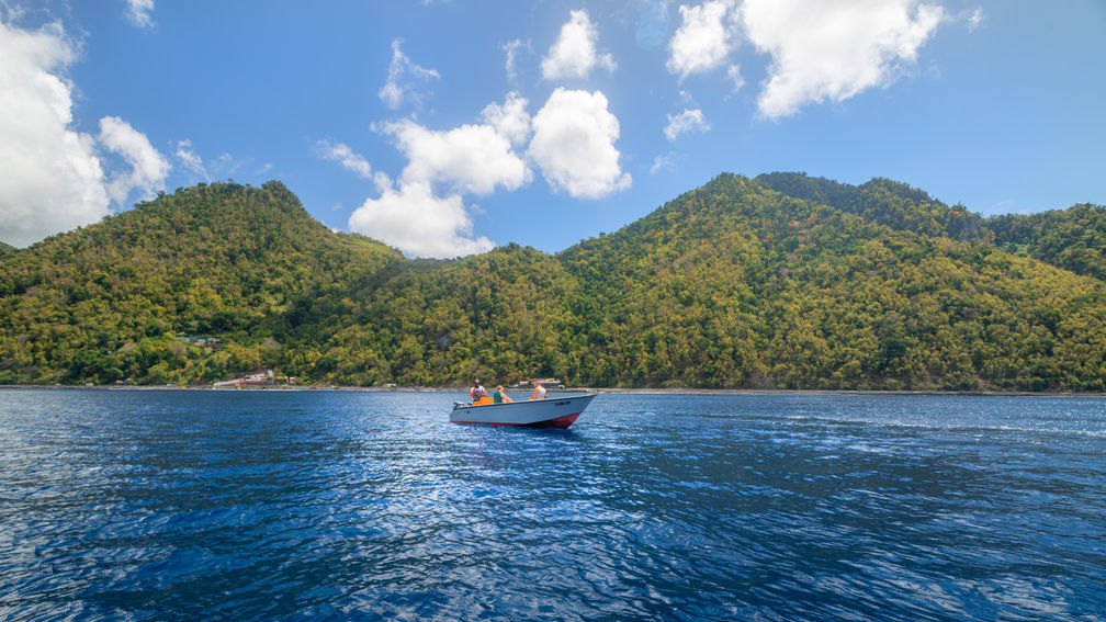 White boat with a red hull floats on blue water near forested hills.